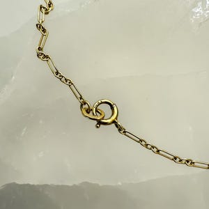 May include: Close-up of a gold-colored necklace with a delicate chain and a spring ring clasp. The chain links are rectangular and connected. The clasp is round with a small lever. The background is a light, textured surface.