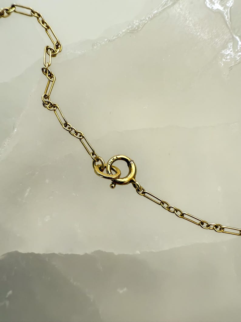 May include: Close-up of a gold-colored necklace with a delicate chain and a spring ring clasp. The chain links are rectangular and connected. The clasp is round with a small lever. The background is a light, textured surface.