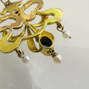 May include: Close-up of a gold pendant with an elaborate, flowing design. It has a dark blue oval gemstone and three small, dangling pearls. The metal has a warm, antique finish, suggesting age and craftsmanship.