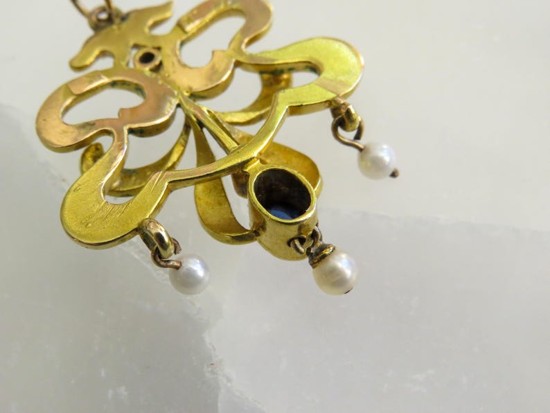 May include: Close-up of a gold pendant with an elaborate, flowing design. It has a dark blue oval gemstone and three small, dangling pearls. The metal has a warm, antique finish, suggesting age and craftsmanship.