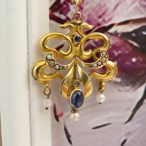 May include: An elaborate gold pendant with a floral design, featuring a central oval blue gemstone and small pearls. The pendant hangs from a delicate gold chain. The background is a soft, blurred floral pattern.