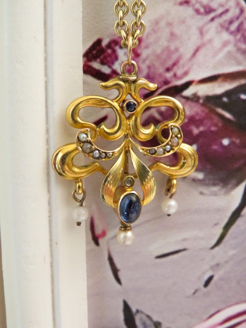 May include: An elaborate gold pendant with a floral design, featuring a central oval blue gemstone and small pearls. The pendant hangs from a delicate gold chain. The background is a soft, blurred floral pattern.