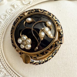 May include: A vintage round brooch with a black center, gold filigree, and pearl accents. The brooch features a floral design with a flower and leaves, all set within a gold frame. The brooch is approximately 2 inches in diameter.