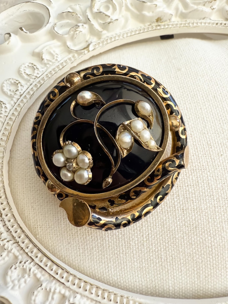May include: A vintage round brooch with a black center, gold filigree, and pearl accents. The brooch features a floral design with a flower and leaves, all set within a gold frame. The brooch is approximately 2 inches in diameter.