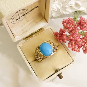 May include: A gold-toned ring with a large, oval turquoise stone, displayed in an open vintage jewelry box. The ring has an intricate filigree design around the stone. A sprig of pink flowers is next to the box.