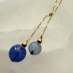 May include: A gold-toned necklace with two dangling strands. Each strand ends with a blue glass orb, one a deep blue and the other a lighter blue. The orbs are accented with gold-colored beads.