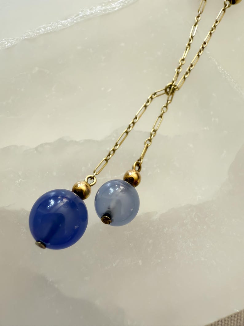 May include: A gold-toned necklace with two dangling strands. Each strand ends with a blue glass orb, one a deep blue and the other a lighter blue. The orbs are accented with gold-colored beads.