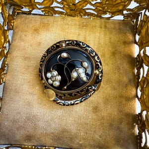 May include: An antique oval brooch with a black center, adorned with pearl accents and gold-toned metal detailing. The brooch features a floral design and is set within a gold-colored decorative box.