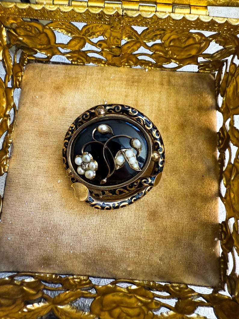 May include: An antique oval brooch with a black center, adorned with pearl accents and gold-toned metal detailing. The brooch features a floral design and is set within a gold-colored decorative box.