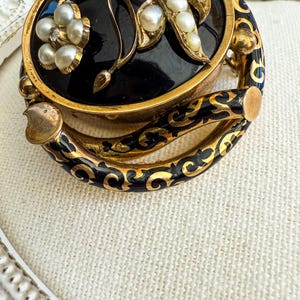 May include: An antique gold-toned brooch with a black enamel center, adorned with pearls and floral designs. The brooch features a hinged design with intricate black and gold enamel detailing around the edges, showcasing a vintage aesthetic.