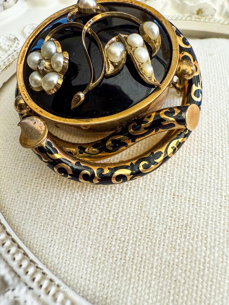 May include: An antique gold-toned brooch with a black enamel center, adorned with pearls and floral designs. The brooch features a hinged design with intricate black and gold enamel detailing around the edges, showcasing a vintage aesthetic.