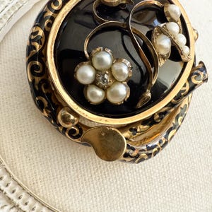 May include: A vintage black and gold brooch with a floral design. The brooch features a central black onyx stone, surrounded by gold filigree and adorned with faux pearls. The floral design includes a flower with a central rhinestone.