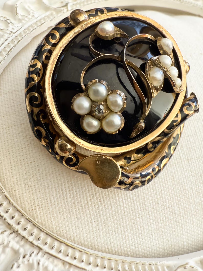 May include: A vintage black and gold brooch with a floral design. The brooch features a central black onyx stone, surrounded by gold filigree and adorned with faux pearls. The floral design includes a flower with a central rhinestone.