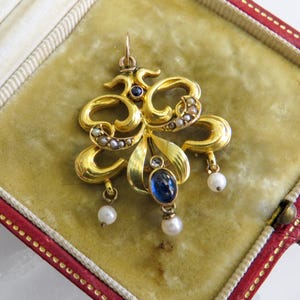 May include: A gold pendant with an elaborate, floral design. It features a large, oval blue gemstone, small pearls, and a small blue gemstone. The pendant is displayed in a red and gold jewelry box.