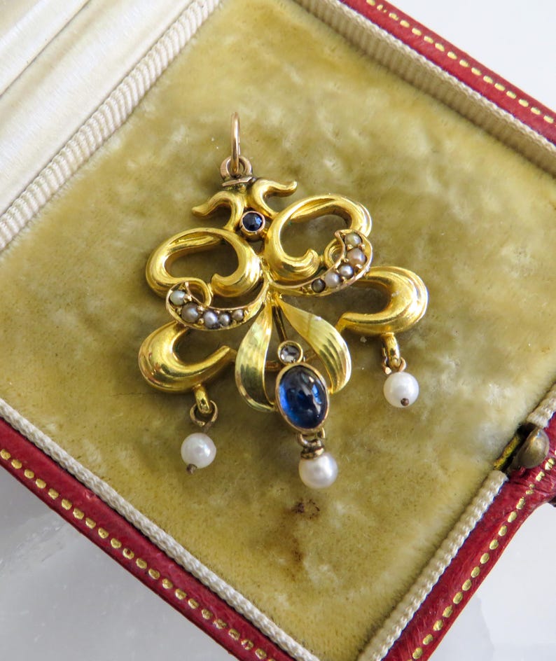 May include: A gold pendant with an elaborate, floral design. It features a large, oval blue gemstone, small pearls, and a small blue gemstone. The pendant is displayed in a red and gold jewelry box.