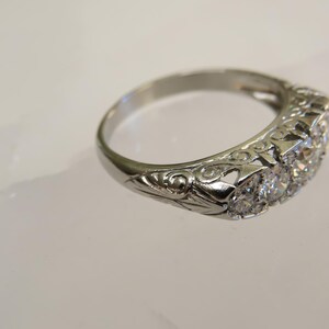 Op de afbeelding: Een zilveren ring met een rij heldere edelstenen. De ring heeft een sierlijk, gegraveerd ontwerp op de band. De edelstenen zijn in een rij geplaatst, de ring ligt op een witte ondergrond.