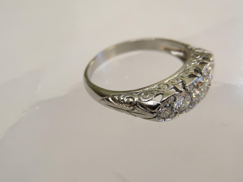 Op de afbeelding: Een zilveren ring met een rij heldere edelstenen. De ring heeft een sierlijk, gegraveerd ontwerp op de band. De edelstenen zijn in een rij geplaatst, de ring ligt op een witte ondergrond.