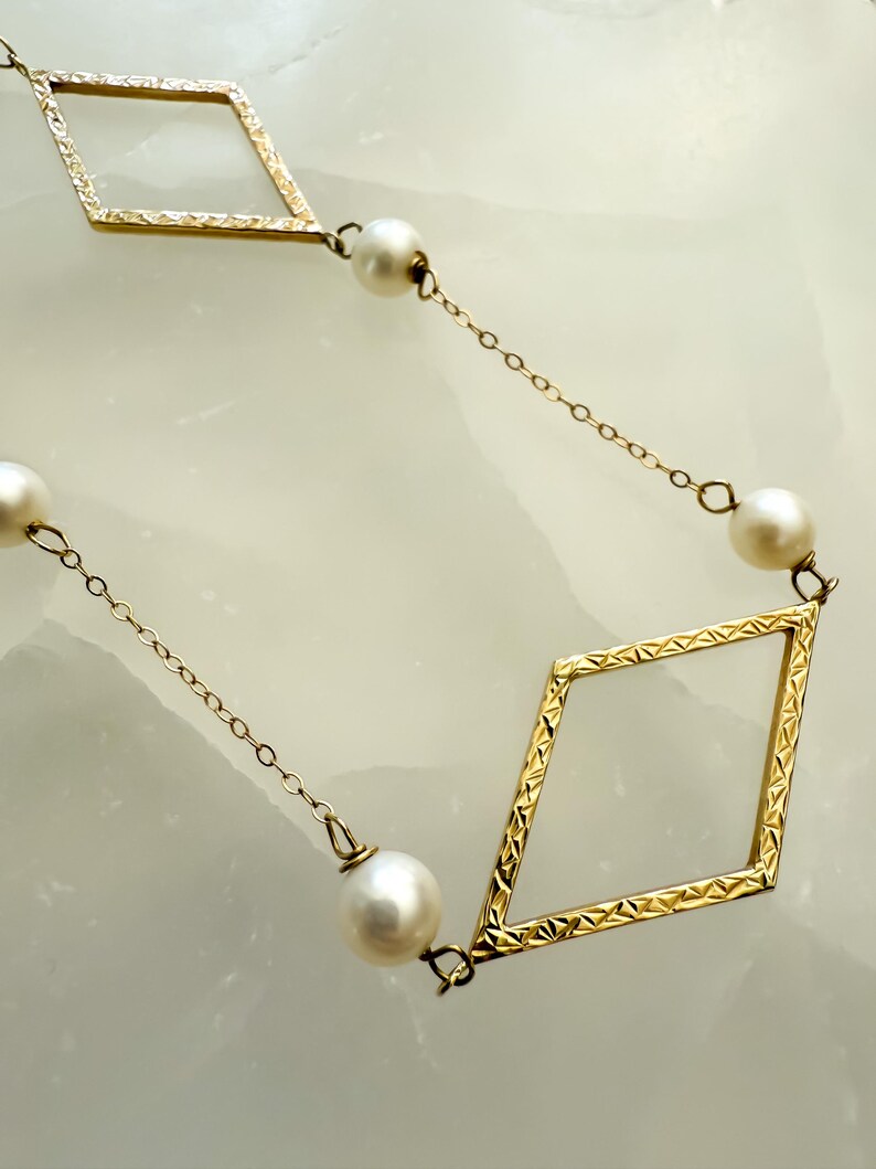 May include: A gold-colored necklace featuring a repeating pattern of diamond-shaped frames and round, white pearls. The chain is delicate, and the diamonds have a textured surface. The pearls are evenly spaced.