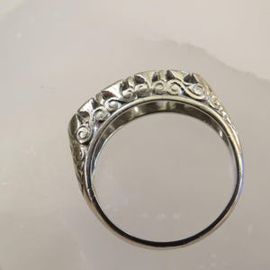 Op de afbeelding: Een zilveren ring met een sierlijk, opgerold ontwerp. De band van de ring is glad, terwijl het bovenste gedeelte gedetailleerde, wervelende patronen heeft. De ring wordt getoond tegen een lichte achtergrond, die de ingewikkelde details en de metalen glans benadrukt.