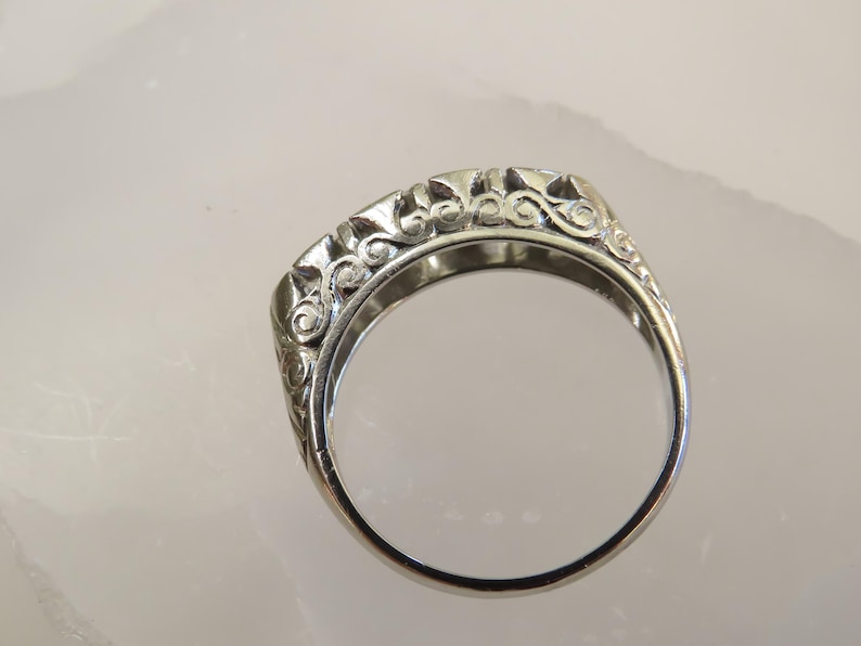 Op de afbeelding: Een zilveren ring met een sierlijk, opgerold ontwerp. De band van de ring is glad, terwijl het bovenste gedeelte gedetailleerde, wervelende patronen heeft. De ring wordt getoond tegen een lichte achtergrond, die de ingewikkelde details en de metalen glans benadrukt.