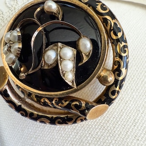May include: Close-up of a vintage brooch with a black enamel center and gold-tone filigree detailing. The brooch features several creamy white pearls and a decorative border with black and gold accents. The design is ornate and elegant.