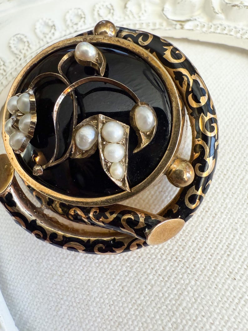 May include: Close-up of a vintage brooch with a black enamel center and gold-tone filigree detailing. The brooch features several creamy white pearls and a decorative border with black and gold accents. The design is ornate and elegant.