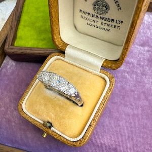 Op de afbeelding: Een vintage diamanten ring gepresenteerd in een bruinleren juwelendoosje met een gele fluwelen binnenkant. De ring heeft een rij sprankelende diamanten. De doos is gemarkeerd met "MAPPIN & WEBB LTD REGENT STREET, LONDON, W."