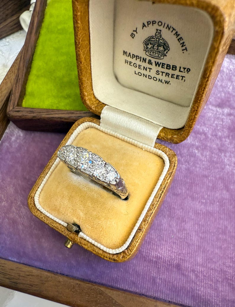 Op de afbeelding: Een vintage diamanten ring gepresenteerd in een bruinleren juwelendoosje met een gele fluwelen binnenkant. De ring heeft een rij sprankelende diamanten. De doos is gemarkeerd met "MAPPIN & WEBB LTD REGENT STREET, LONDON, W."
