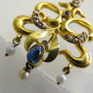 May include: Close-up of a gold-toned pendant with an ornate floral design. The pendant features a large oval blue gemstone, small pearls, and tiny clear stones. Two dangling pearls hang from the bottom.