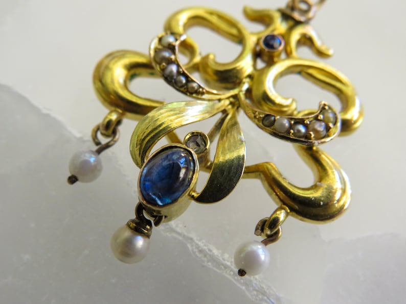 May include: Close-up of a gold-toned pendant with an ornate floral design. The pendant features a large oval blue gemstone, small pearls, and tiny clear stones. Two dangling pearls hang from the bottom.