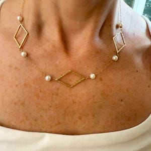 May include: A gold-colored necklace featuring a delicate chain with diamond-shaped pendants and small, round pearls. The necklace is worn against a white top, showcasing the jewelry's design and details.