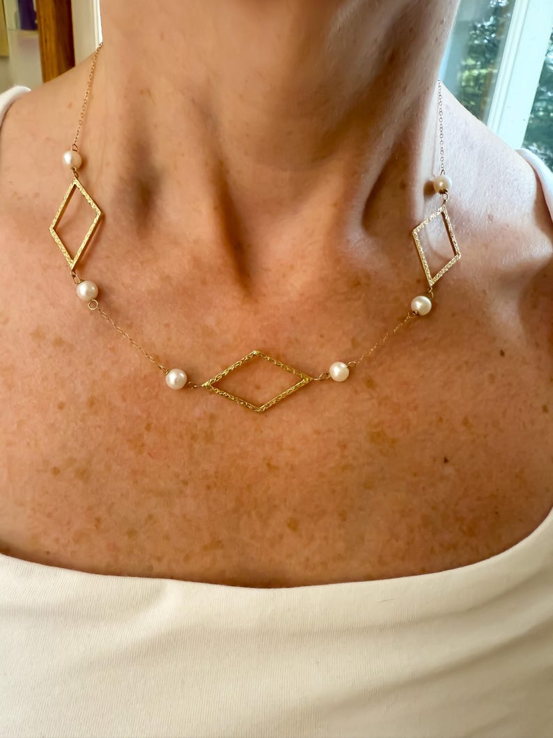 May include: A gold-colored necklace featuring a delicate chain with diamond-shaped pendants and small, round pearls. The necklace is worn against a white top, showcasing the jewelry's design and details.