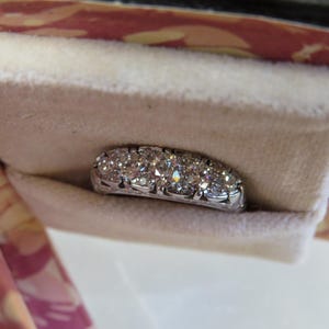 Op de afbeelding: Een sprankelende diamanten ring met meerdere rond geslepen diamanten, gezet in een witte metalen band. De ring wordt gepresenteerd in een zacht, cr&egrave;mekleurig fluwelen zakje, tegen een roze en rode achtergrond met patronen.