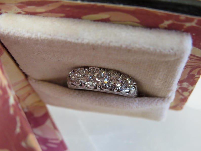 Op de afbeelding: Een sprankelende diamanten ring met meerdere rond geslepen diamanten, gezet in een witte metalen band. De ring wordt gepresenteerd in een zacht, cr&egrave;mekleurig fluwelen zakje, tegen een roze en rode achtergrond met patronen.
