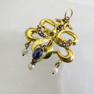May include: A gold pendant with an elaborate, swirling design. It features a large oval blue gemstone, small pearls, and tiny seed pearls. The pendant has a loop at the top for a chain.