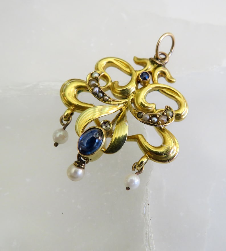 May include: A gold pendant with an elaborate, swirling design. It features a large oval blue gemstone, small pearls, and tiny seed pearls. The pendant has a loop at the top for a chain.