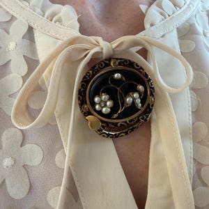 May include: An oval, vintage brooch with a black center, gold filigree border, and floral design. The brooch features small white pearls and a gold-colored leaf accent. It is pinned to a cream-colored blouse with a bow.