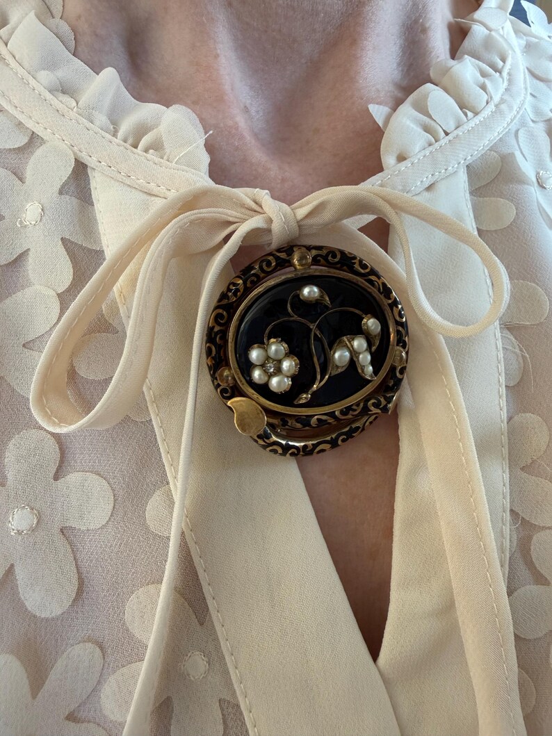 May include: An oval, vintage brooch with a black center, gold filigree border, and floral design. The brooch features small white pearls and a gold-colored leaf accent. It is pinned to a cream-colored blouse with a bow.