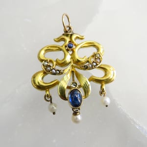 May include: A gold pendant with an elaborate design. It features a central blue gemstone and small pearls. The pendant has a loop at the top for hanging and three dangling pearls. The design includes curved lines and floral elements.