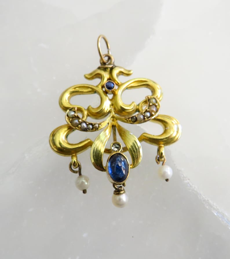 May include: A gold pendant with an elaborate design. It features a central blue gemstone and small pearls. The pendant has a loop at the top for hanging and three dangling pearls. The design includes curved lines and floral elements.