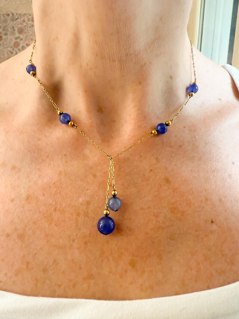 May include: A delicate gold necklace featuring a series of deep blue beads interspersed with smaller gold beads. The necklace has a Y-shaped drop with three additional blue beads. The necklace is worn around the neck.