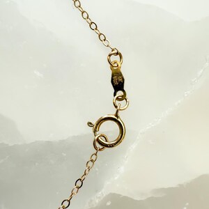 May include: Close-up of a gold-colored necklace chain with a spring ring clasp. The chain is made of small, delicate links. The clasp is attached to a small tag with stamped markings. The background is a light, marbled surface.