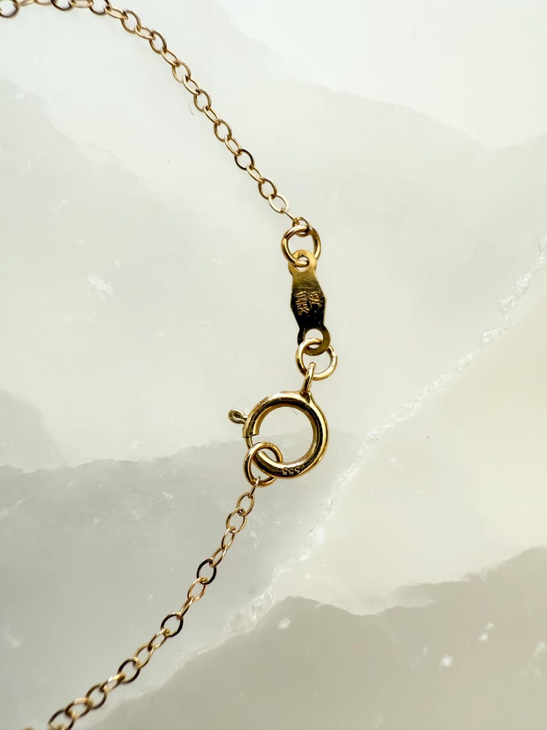 May include: Close-up of a gold-colored necklace chain with a spring ring clasp. The chain is made of small, delicate links. The clasp is attached to a small tag with stamped markings. The background is a light, marbled surface.