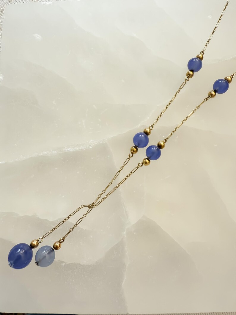 May include: A delicate necklace featuring a gold chain with alternating blue beads and gold accents. The necklace has a Y-shaped design, with larger blue beads at the bottom. The beads are a soft, muted blue color.