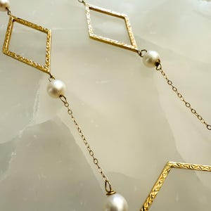 May include: Gold-toned necklace featuring diamond-shaped links with textured detailing, interspersed with round, white pearls. The necklace is displayed against a light-colored background, highlighting the contrast between the gold and pearls.