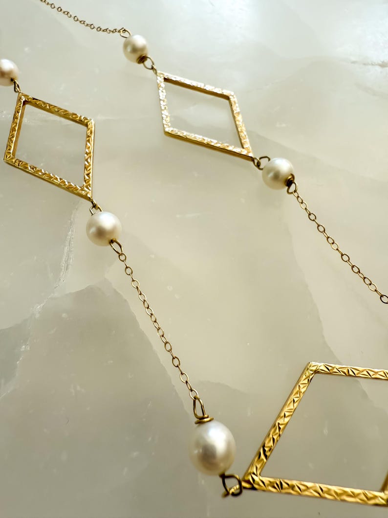 May include: Gold-toned necklace featuring diamond-shaped links with textured detailing, interspersed with round, white pearls. The necklace is displayed against a light-colored background, highlighting the contrast between the gold and pearls.