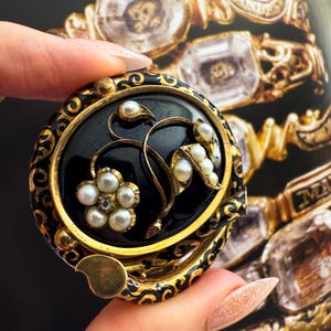 May include: A round, black and gold brooch with a floral design. The brooch features a black center with a gold border and accents. The floral design includes small pearls and gold detailing. The brooch is held in a hand.