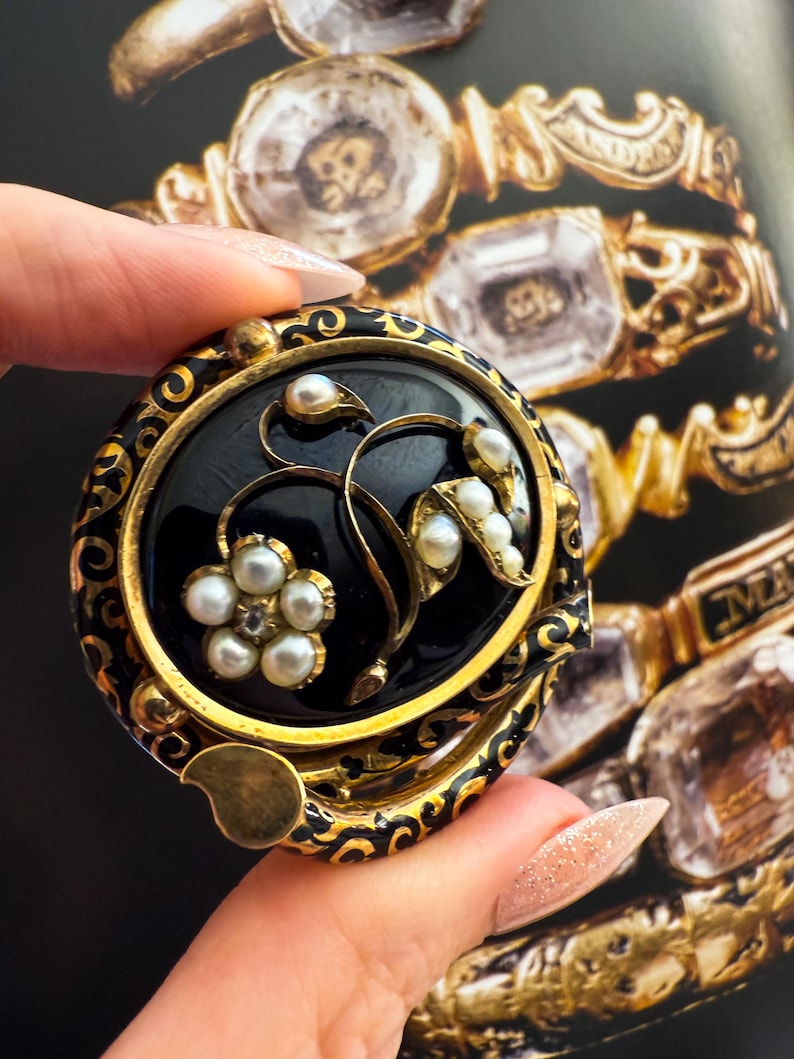 May include: A round, black and gold brooch with a floral design. The brooch features a black center with a gold border and accents. The floral design includes small pearls and gold detailing. The brooch is held in a hand.