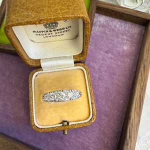 Op de afbeelding: Een antieke ring met meerdere kleine, heldere stenen in een zilveren band, gepresenteerd in een open, antieke leren sieradendoos. De doos heeft een gele fluwelen binnenkant en de tekst "MAPPIN & WEBB LTD REGENT STREET, LONDON,W."