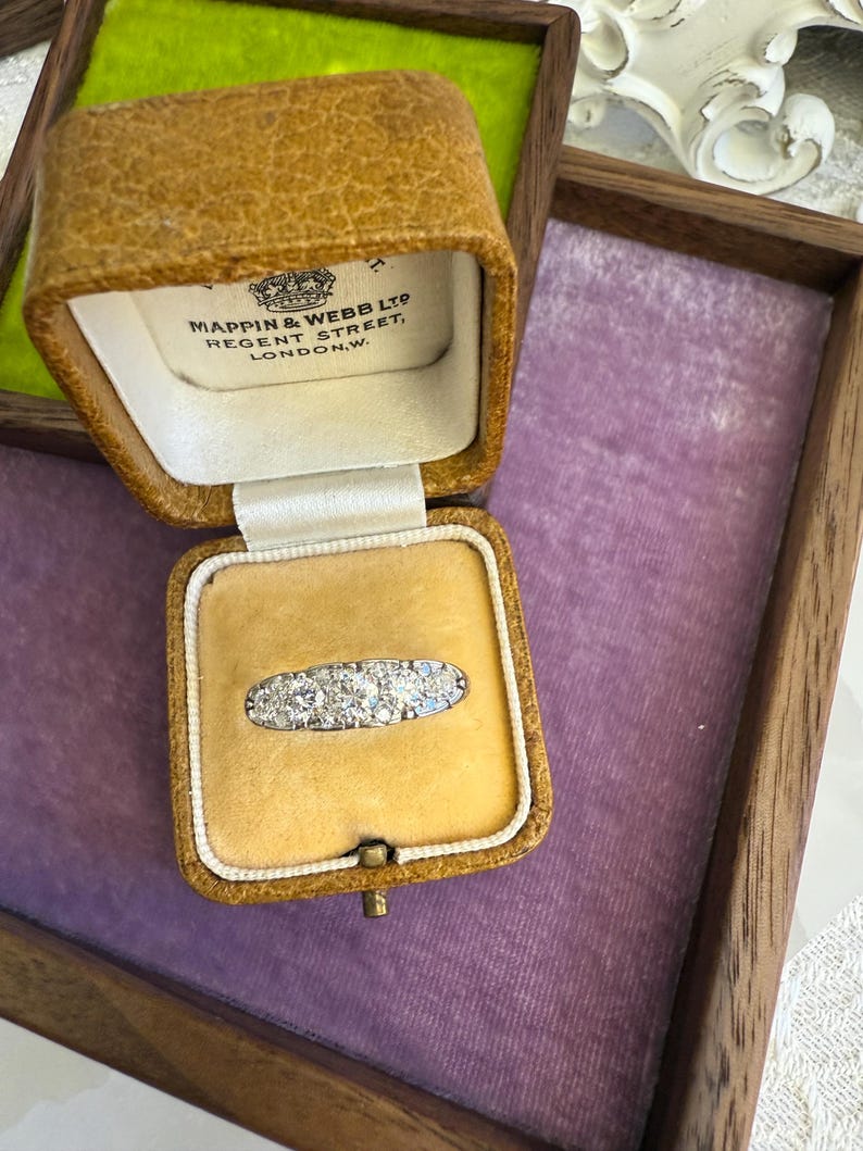 Op de afbeelding: Een antieke ring met meerdere kleine, heldere stenen in een zilveren band, gepresenteerd in een open, antieke leren sieradendoos. De doos heeft een gele fluwelen binnenkant en de tekst "MAPPIN & WEBB LTD REGENT STREET, LONDON,W."